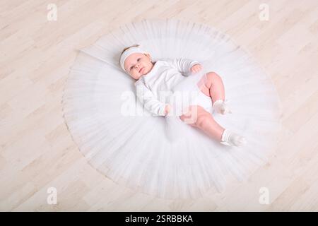 Infant female with light hair, lying on wooden floor in full white tutu. Expression calm and curious. Soft lighting enhances serene setting Stock Photo