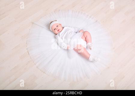 Infant female with light hair, lying on wooden floor in full white tutu. Expression calm and curious. Soft lighting enhances serene setting Stock Photo