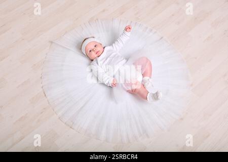 Infant female with light hair, lying on wooden floor in full white tutu. Expression calm and curious. Soft lighting enhances serene setting Stock Photo