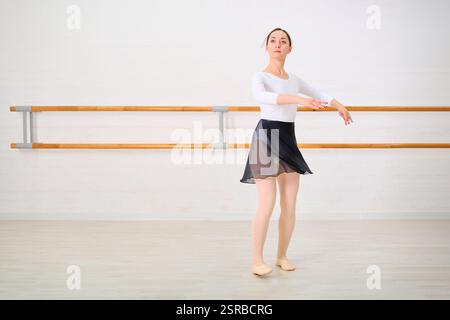 Young female ballerina with brown hair practicing in dance studio. Standing on pointe with focused expression. Bright lighting against simple, white b Stock Photo
