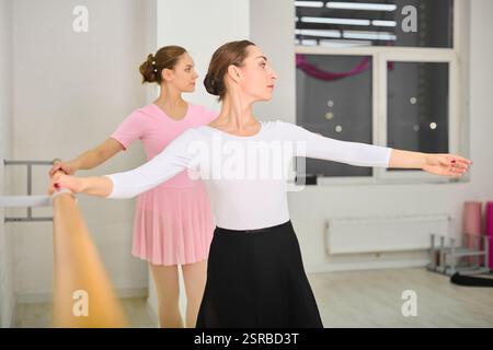Two young adult female ballerinas with brown hair practice at ballet barre. They perform graceful moves in bright studio with neutral walls and ample Stock Photo