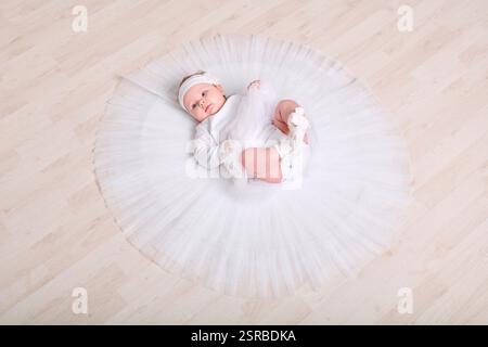 Infant female with light hair, lying on wooden floor in full white tutu. Expression calm and curious. Soft lighting enhances serene setting Stock Photo
