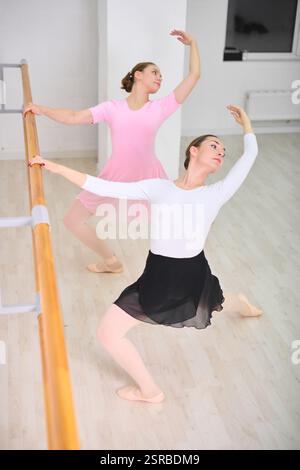 Two young female ballerinas in pink and white dance outfits practice at ballet barre. Wooden floor and white walls create bright studio atmosphere. Gr Stock Photo