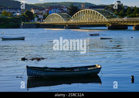 Boats floating on a calm river beneath an arched bridge in a peaceful town setting. Colindres, cantabria, spain Stock Photo