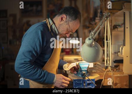 Concentrated italian luthier carefully working on a new violin scroll secured in a blue vise, using precise tools under a warm lamp light in his works Stock Photo