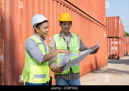 Engineers analyzing logistics plans in a container yard, focusing on project management and safety. Stock Photo