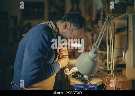 Italian luthier is accurately working with a gouge on the scroll of a new violin in his workshop, illuminated by a lamp, with care and concentration Stock Photo