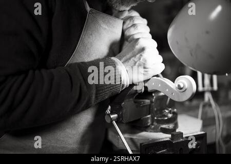 Craftsman deeply engaged in woodworking with a lamp on a workbench, shaping the peg box of a violin scroll Stock Photo