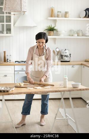 Confident mature woman flattening dough with rolling pin in kitchen Stock Photo