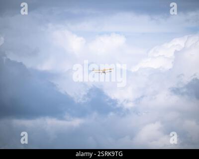 A small yellow biplane soars high in the sky, surrounded by dramatic white and gray clouds. The vast sky emphasizes the sense of adventure and freedom Stock Photo