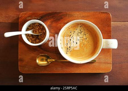 A cup of frothy hot coffee with brown sugar pot and brass teaspoon on wooden table Stock Photo