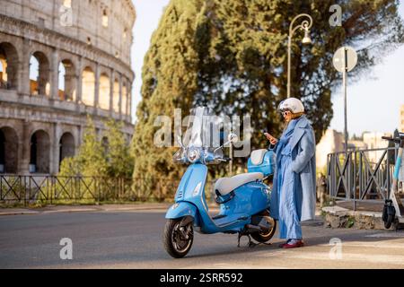 A fashionable woman in a blue coat and jumpsuit stands by her scooter, checking her phone on a sunny day in Rome, Italy. A modern take on urban elegance, independence, and city lifestyle Stock Photo