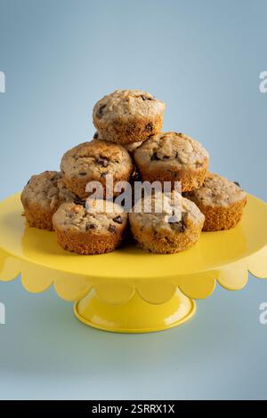 A stack of banana and chocolate chip muffins, beautifully arranged on a vibrant yellow cake stand against a soft blue background Stock Photo