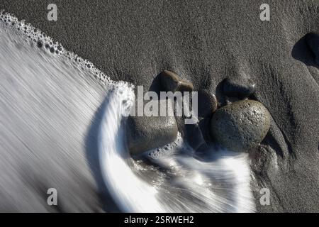 A slow shutter speed captures ocean waves flowing over black sand and smooth stones on a serene beach Stock Photo