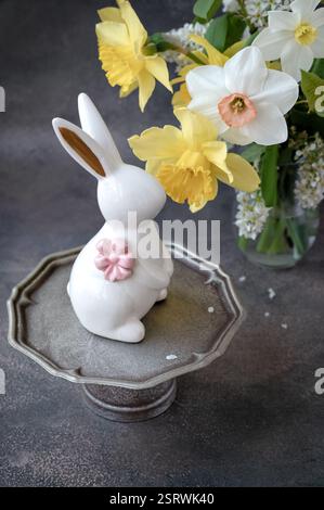 A delightful rabbit figurine adorned with a pink flower sits on a metallic stand, surrounded by stunning yellow daffodils and white blossoms in a cozy Stock Photo