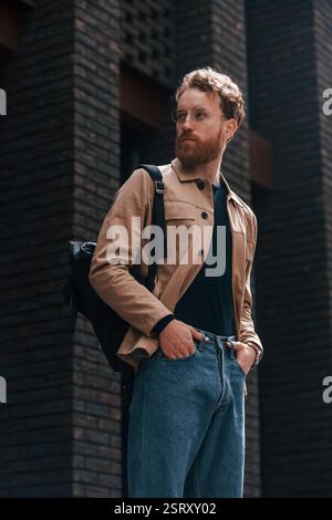 Stylish man with beard in khaki colored jacket and in jeans is outdoors near building. Stock Photo