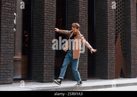 Having fun by walking the sidewalk. Stylish man with beard in khaki colored jacket and in jeans is outdoors near building. Stock Photo