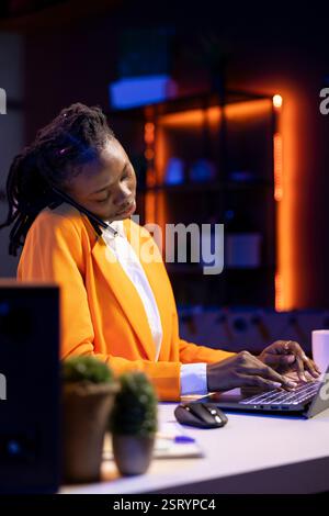 Student at desk talking with school colleague in phone call while using notebook to research information online for project. Girl doing telephone call with friend while doing homework on laptop Stock Photo