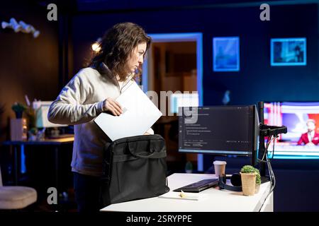 Woman packing laptop after finishing doing software quality assurance and reading source code. Engineer placing notebook in backpack after inspecting coding on desktop PC and finding all issues Stock Photo