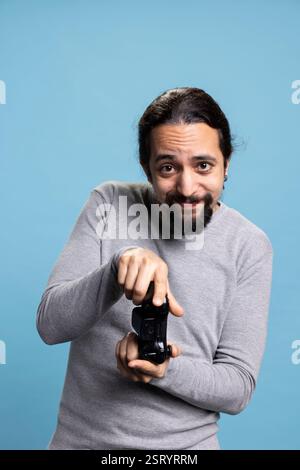Middle eastern man playing video games online on his console, attending an electronic cyber tournament against blue background. Young adult having fun with a shooter in studio, controller. Stock Photo