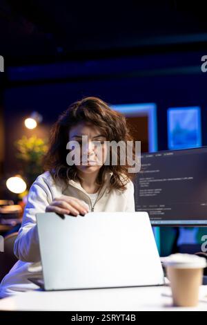 IT expert working from home, opening laptop, testing and deploying programs and systems. Engineer checking code displayed on computer monitors, sitting at desk chair in apartment Stock Photo