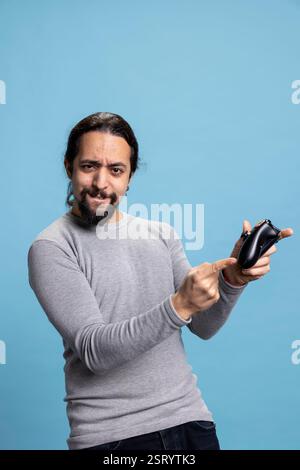 Overjoyed young adult winning an online tournament in the studio, feeling pleased and proud of his victory against blue background. Satisfied guy smiling in front of camera, video games on console. Stock Photo