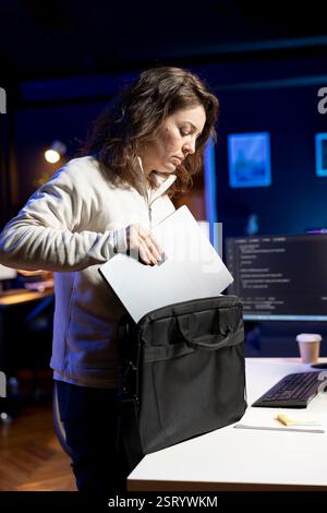Technician packing laptop at end of work shift after securing company servers from virus attacks. Woman putting notebook in backpack, leaving office after building firewalls protecting data Stock Photo