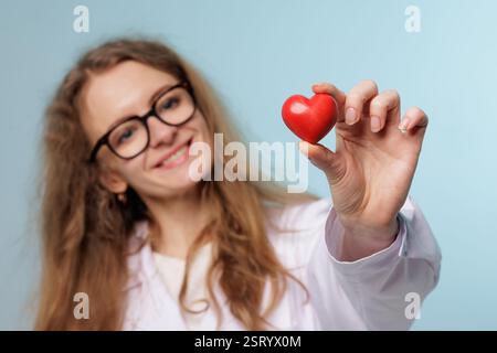 Smiling female doctor holding a red heart, symbolizing cardiology, healthcare, heart health, and medical care on a light blue background. Stock Photo