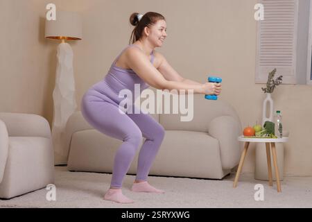Smiling overweight woman in sportswear doing squats with dumbbells at home promoting fitness, weight loss and healthy lifestyle in cozy interior. Stock Photo