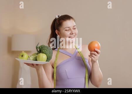 Smiling overweight woman in tracksuit holding healthy fruits and vegetables promoting balanced diet, weight loss and healthy lifestyle. Stock Photo