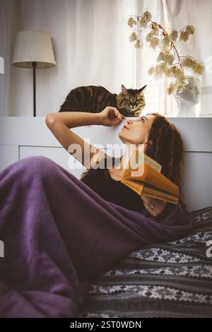 Woman is relaxing at home. Lying in bed with a book and a cat Stock Photo