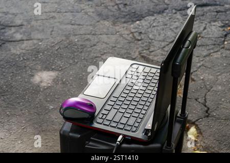 A laptop is placed on a suitcase with a purple mouse beside it. The setting features a textured asphalt surface, indicative of an outdoor workspace. S Stock Photo