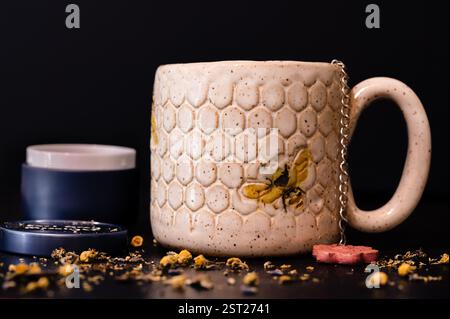A handmade ceramic mug surrounded by loose chamomile tea and a blue tin in the background Stock Photo