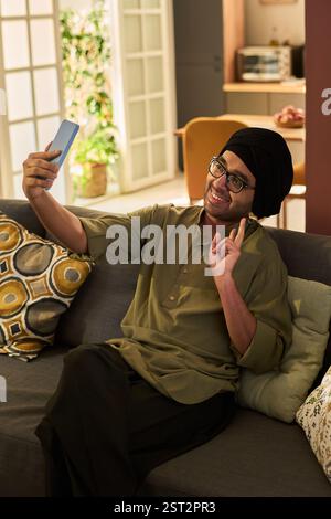 Man wearing casual outfit sitting on couch and smiling while taking selfie with smartphone in bright living room holding cup engaging in leisure activity Stock Photo