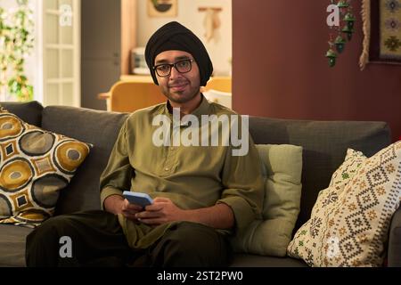 Portrait of young man sitting on couch in cozy living room holding smartphone, wearing turban and glasses with green shirt, offering warm smile and relaxed demeanor Stock Photo