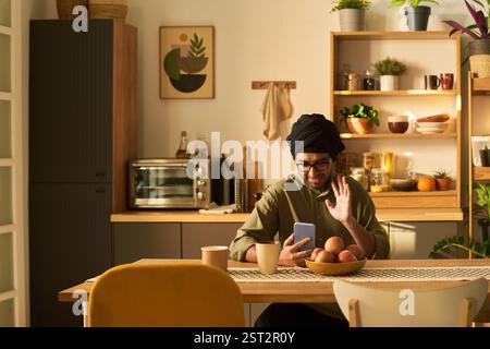 Man sitting in cozy kitchen, holding smartphone while touching beard and sitting at table with bowl of fruit. Warm ambient lighting highlighting kitchen decor and wooden elements Stock Photo