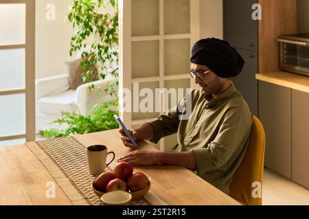 Young man with turban sitting at kitchen table reading digital tablet while drinking coffee with fruits on table and greenery in background Stock Photo