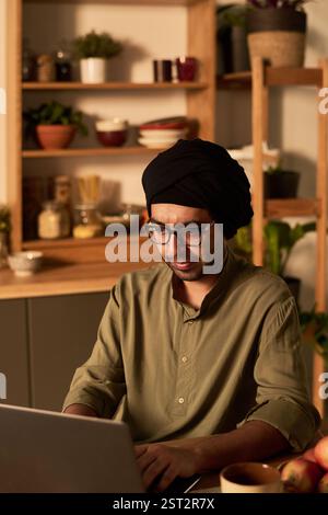 Man sitting in home kitchen with wooden shelves and plants in background, focused on using a laptop for work. Warm ambient lighting enhancing comfortable atmosphere Stock Photo