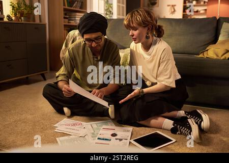 Two people sitting on floor, closely examining financial documents and discussing planning. Papers, forms, tablets scattered around indicating collaborative effort in budgeting Stock Photo