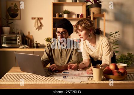 Diverse team working together on laptop while sitting at wooden table in home office atmosphere, reviewing documents and discussing ideas Stock Photo