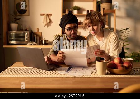 A couple is siting at a kitchen table, reviewing financial documents attentively. Both individuals are focused on paperwork, with a laptop, coffee mugs, and fruit centerpiece visible Stock Photo
