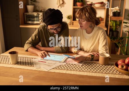 Two individuals sitting at a wooden table, reviewing documents and using a tablet. Kitchen background with plants and decorated shelves, creating a warm and inviting atmosphere Stock Photo