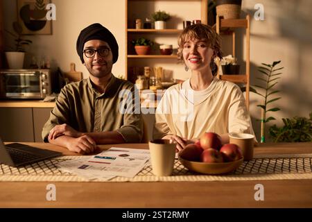 Smiling professionals sitting at office table with documents, laptop, and apples. Comfortable and welcoming workspace with shelves and kitchen appliances in background Stock Photo