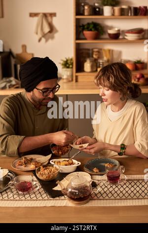 Man and woman sharing a meal in contemporary kitchen setting. Table filled with diverse array of dishes, showcasing a blend of cultural cuisines Stock Photo