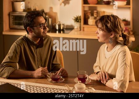Two people sitting at kitchen table, engaged in lively conversation with tea cups in hands. Kitchen environment with modern design elements creating cozy ambiance Stock Photo