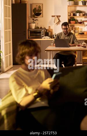 Cozy evening scene with a man using laptop at table in background and a woman using smartphone in foreground. Warm lighting creating inviting atmosphere in modern home setting Stock Photo