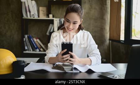 A beautiful, smiling Asian female architect or interior designer using her smartphone at her desk in the office. chatting, responding to messages, scr Stock Photo