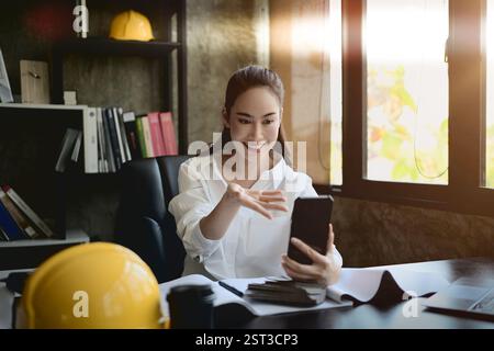 A confident, beautiful Asian female interior designer or architect talking on a video call through her smartphone with a client at her desk in the off Stock Photo