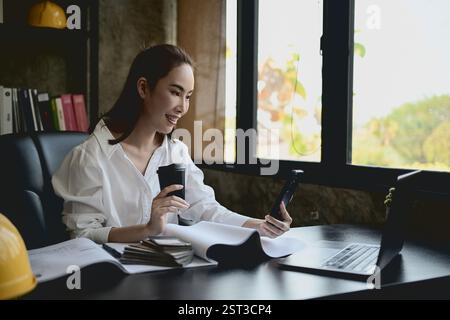 A beautiful, smiling Asian female architect or interior designer using her smartphone at her desk in the office. chatting, responding to messages, scr Stock Photo