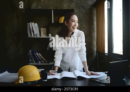 An attractive, confident Asian female interior designer or architect leans over her desk in the office, gazing out the window with a thoughtful expres Stock Photo